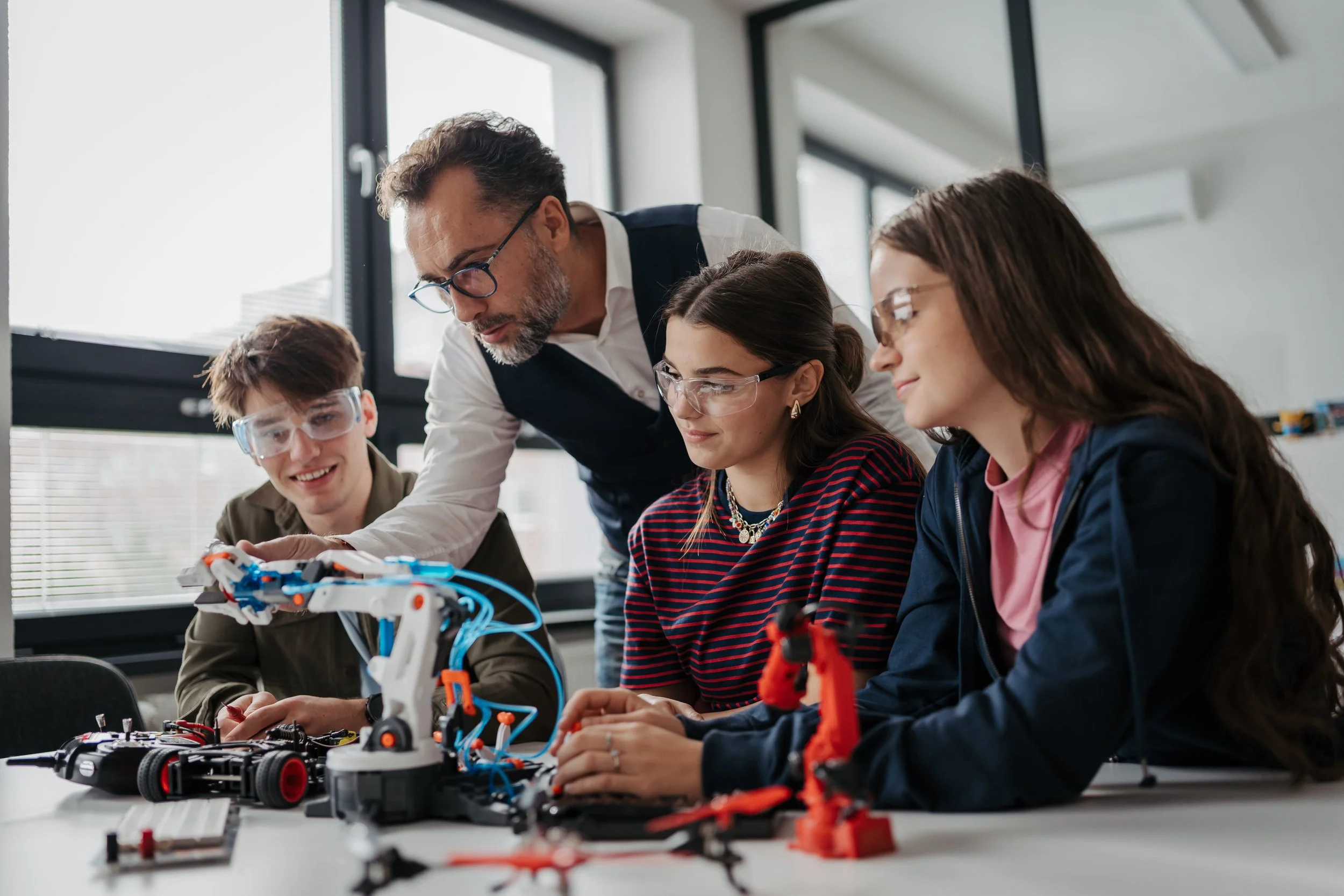 A high school teacher instructs three students on robotics in a classroom, with robotic arms and tools on the table.
