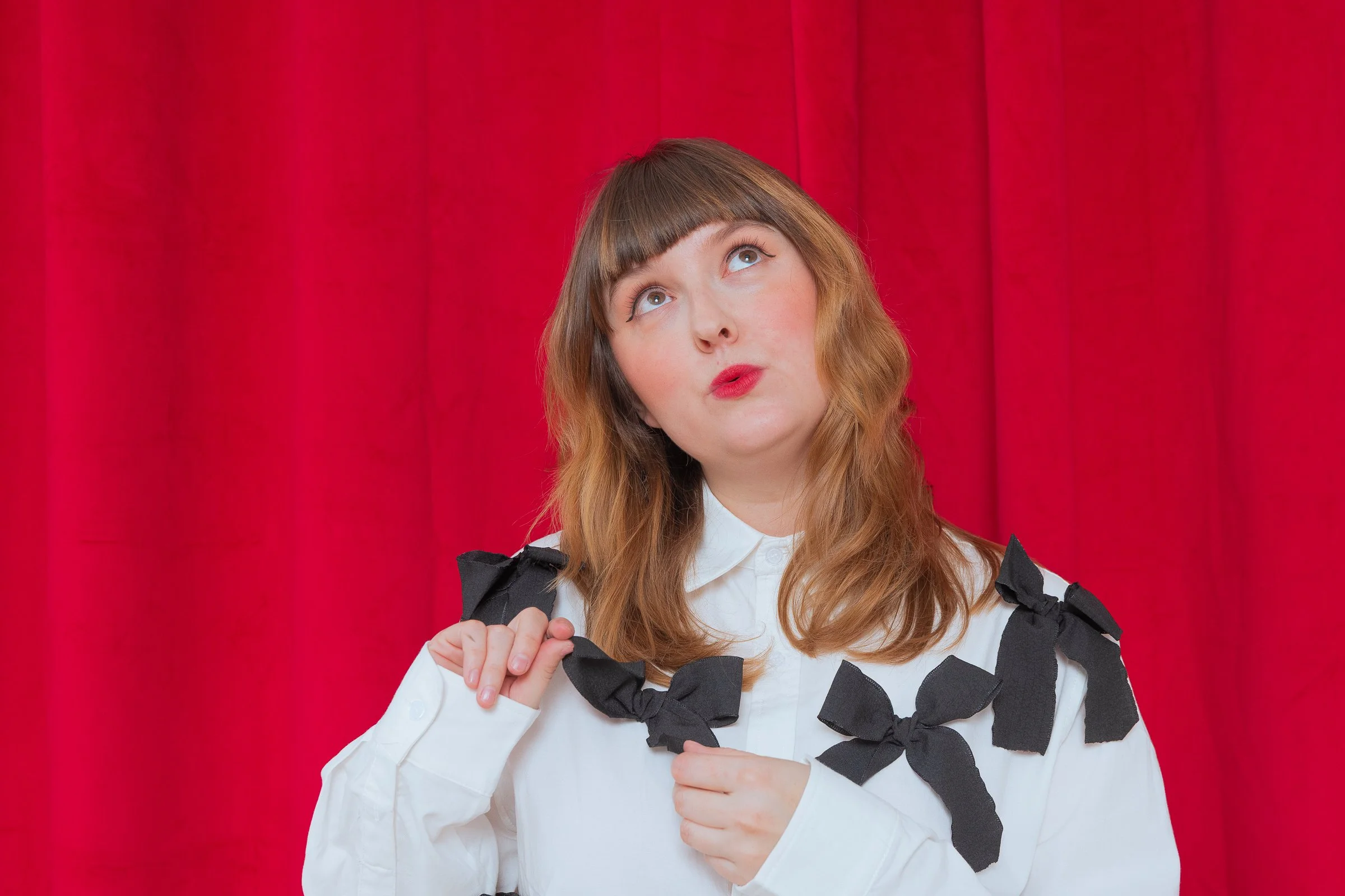 Junge Frau mit mittelbraunen Haaren und roten Lippen trägt ein weißes Hemd mit schwarzen Bändern, steht vor roten Vorhängen und schaut nach oben.