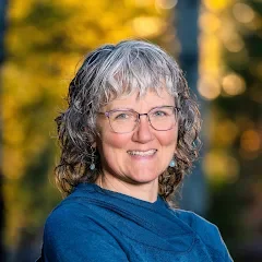 A woman with gray hair, wearing a light blue dress with purple shawl, standing in a green forest, smiling with arms outstretched.