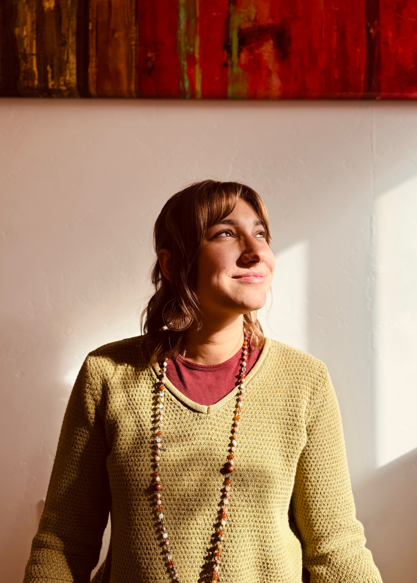 A woman with short dark hair, glasses, and a nose ring smiling with one arm raised behind her head, sitting on a cushion on a wooden floor in a room with plain walls and framed artwork.