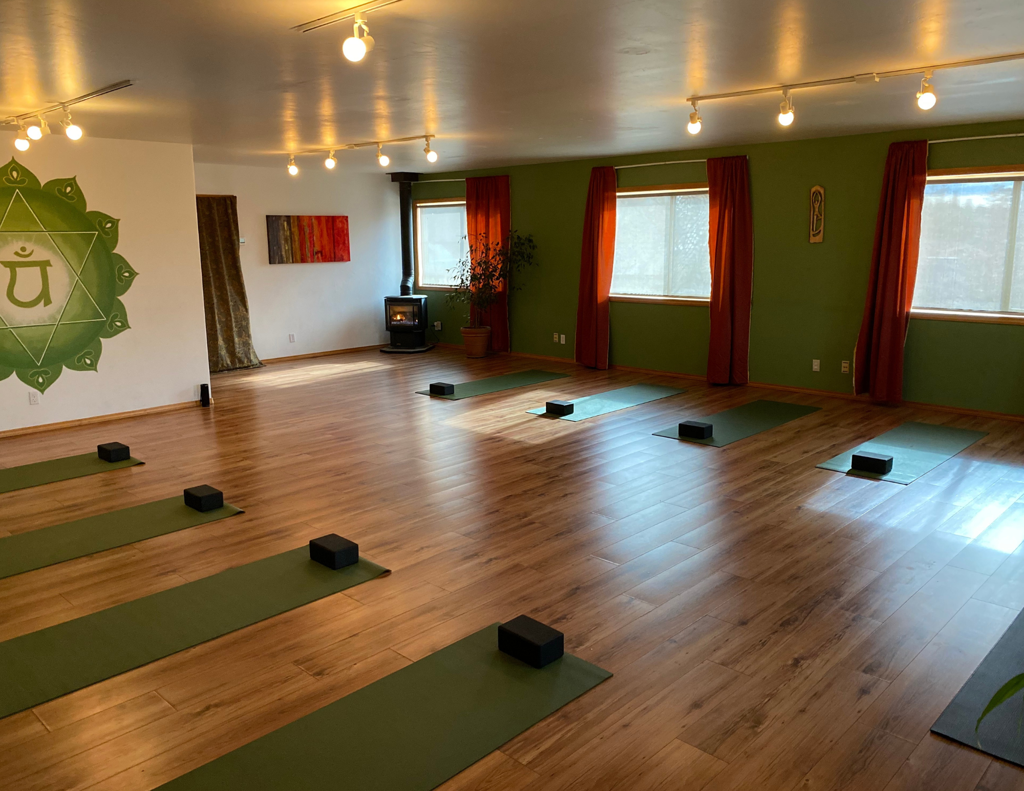 A spacious yoga or meditation room with wooden floors, green walls, and red curtains covering large windows. There is a wall decoration featuring a green circle with a symbol inside, and a small shelf with a statue. The room is lit with ceiling spotlights.
