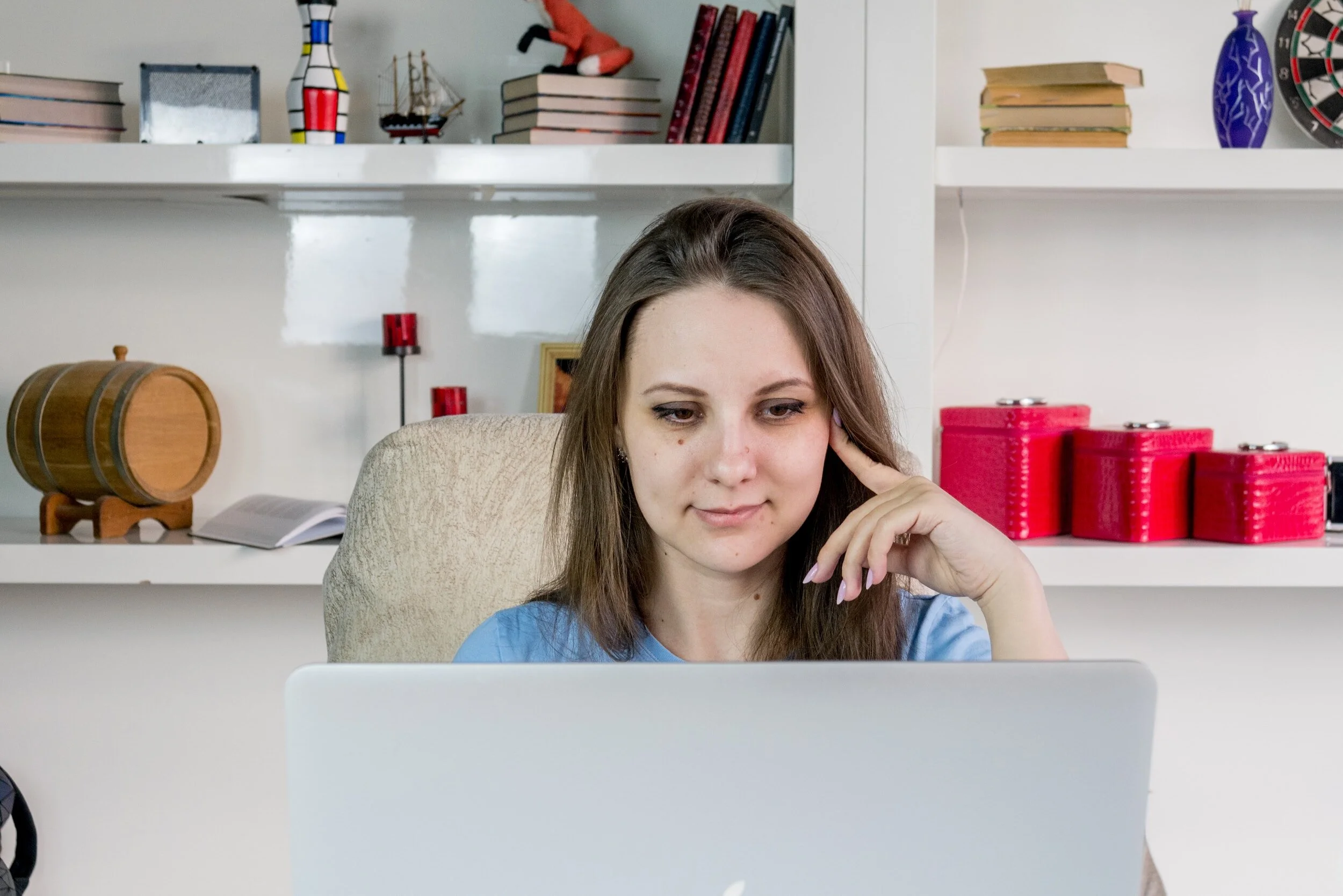 A young woman with brown hair looking at a laptop screen, sitting in a beige chair in front of a white bookshelf with decorative items and red boxes.