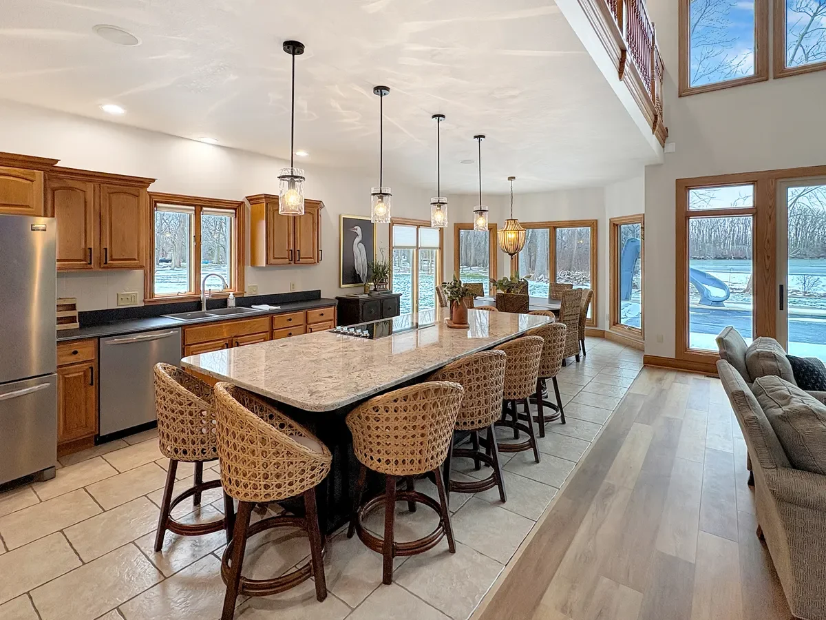 Open-concept kitchen and dining area with large windows overlooking a snowy outdoor landscape, featuring wooden cabinets, a large kitchen island with bar stools, and a seating area with a sliding glass door leading outside.