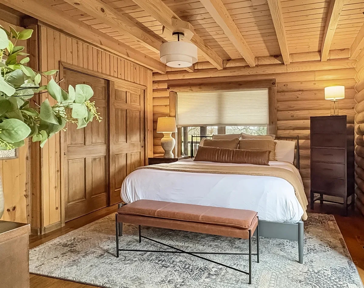 A cozy bedroom with wooden walls and ceiling, a large bed with white and brown bedding, a bench at the foot of the bed, a window with a white blind behind the bed, and two lamps on nightstands on each side of the bed.