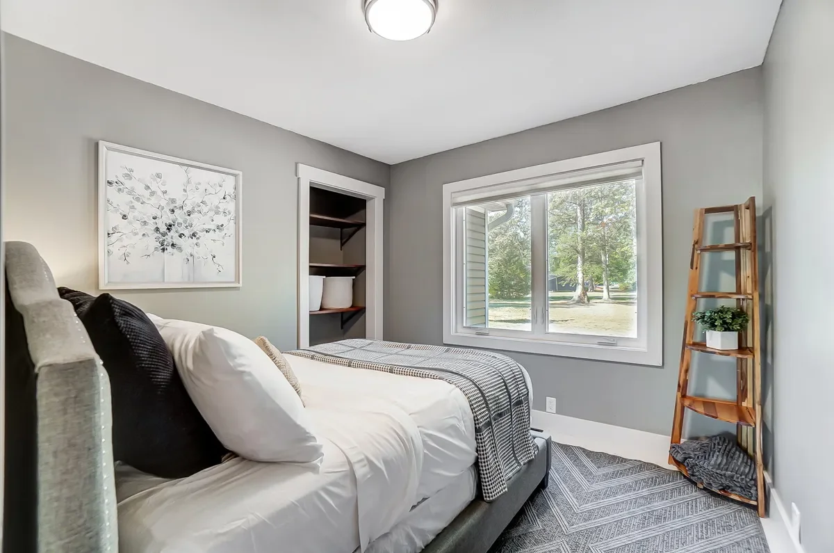 A cozy bedroom with a bed dressed in white bedding and decorative pillows, a window showing a backyard with trees, a wooden ladder shelf with a plant, an art piece on the wall, and neutral-colored walls and carpeting.