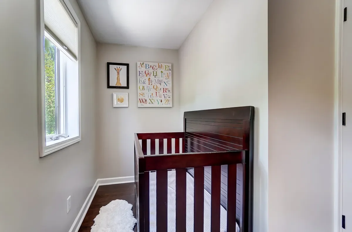 View of a small nursery with a dark wooden crib, white walls, a window with blinds, and framed children's artwork on the wall.