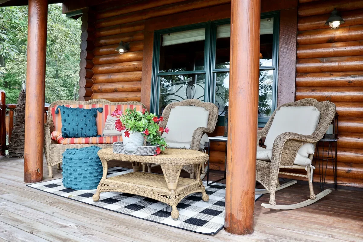 Front porch of a log cabin with wicker furniture, a rocking chair, a small table with a flower arrangement, and decorative cushions, overlooking a wooded area.