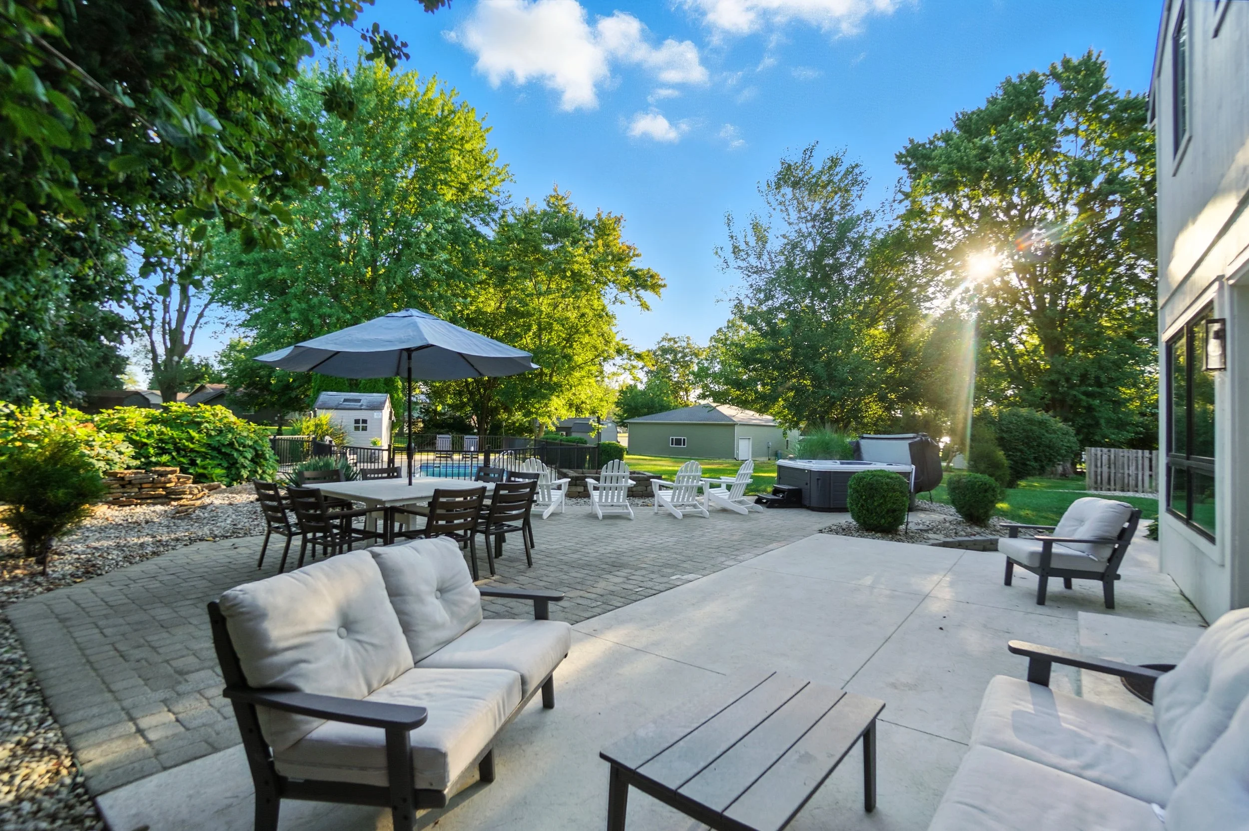 Backyard patio with cushioned outdoor furniture, a dining table with an umbrella, a swimming pool with lounge chairs, trees, and a hot tub, during sunny weather.