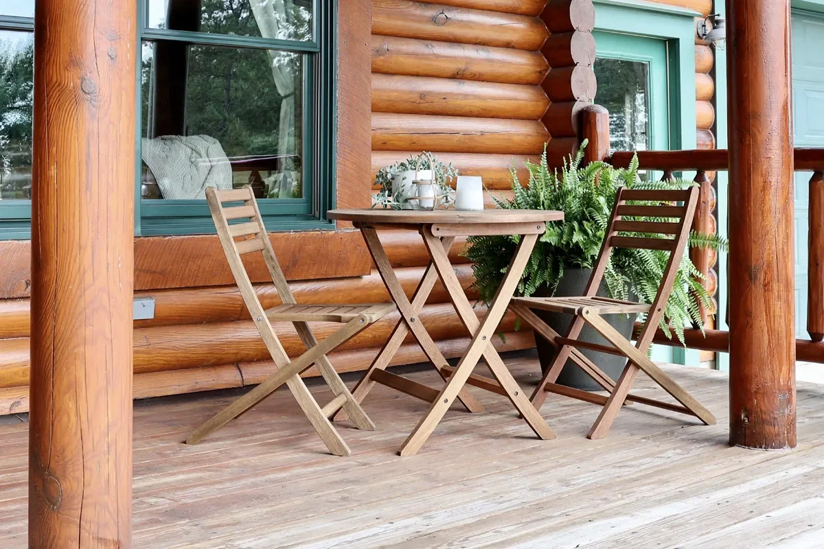 A cozy outdoor patio with a small wooden table set for two, accompanied by matching folding chairs, on a wooden deck outside a log cabin-style home with large windows and green trim, decorated with potted ferns and a small plant.