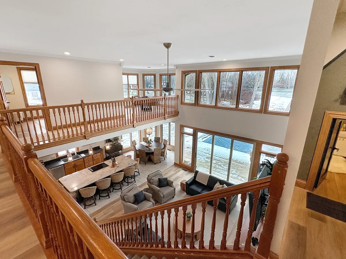 Interior view of a two-story living room, showing a wooden staircase and railing, with large windows overlooking a snowy outdoor scene, a sitting area with sofas, a television, and a kitchen with a dining table.