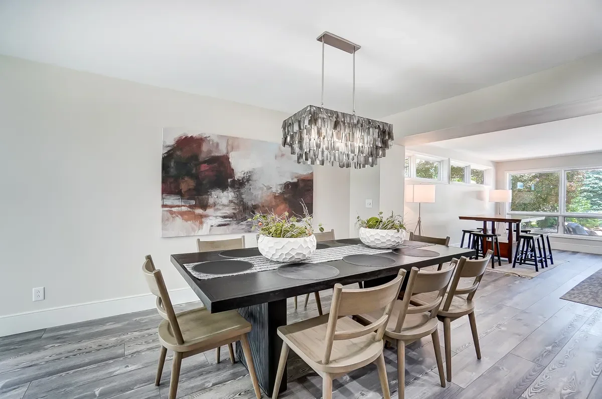 Dining room with black table, light wood chairs, and modern chandelier, artwork on wall, and large windows with natural light.