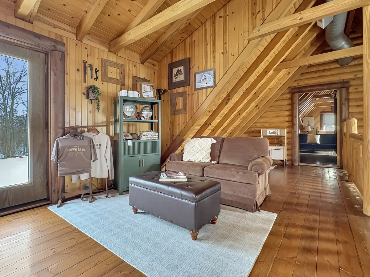 Cozy living room with wood-paneled walls and ceiling, a brown couch with a white pillow, a black ottoman, a green shelf with decor, clothes hanging on a rack, and framed pictures on the wall. There is a large window showing a snowy outdoor scene.