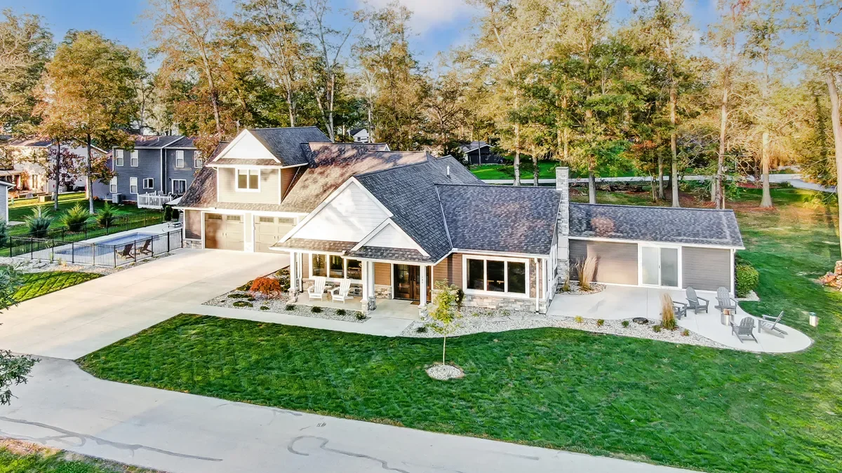 Aerial view of a modern house with a well-maintained lawn, surrounded by tall trees, featuring a front porch, large windows, and a detached garage, with outdoor seating and a small tree in the yard.