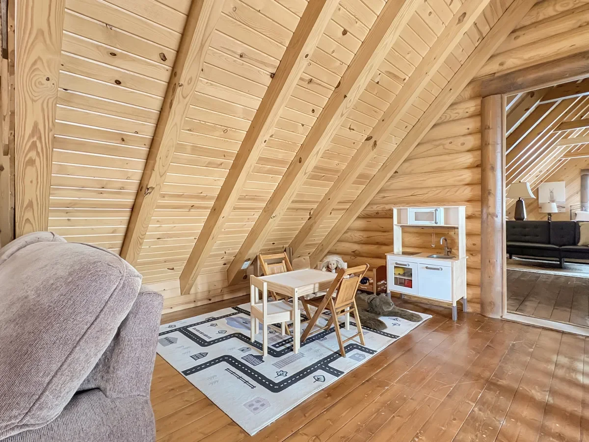 Interior of a cozy wood-paneled attic space with a small children's play area, including a white play kitchen, a table and chairs, and a rug with a road pattern. Part of a gray sofa is visible on the left, and the background shows a living room with 