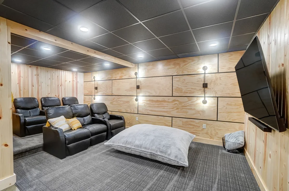 A cozy home theater room with black leather recliners, a large gray floor cushion, a flat-screen TV mounted on the right wall, and wood-paneled walls with modern lighting fixtures.