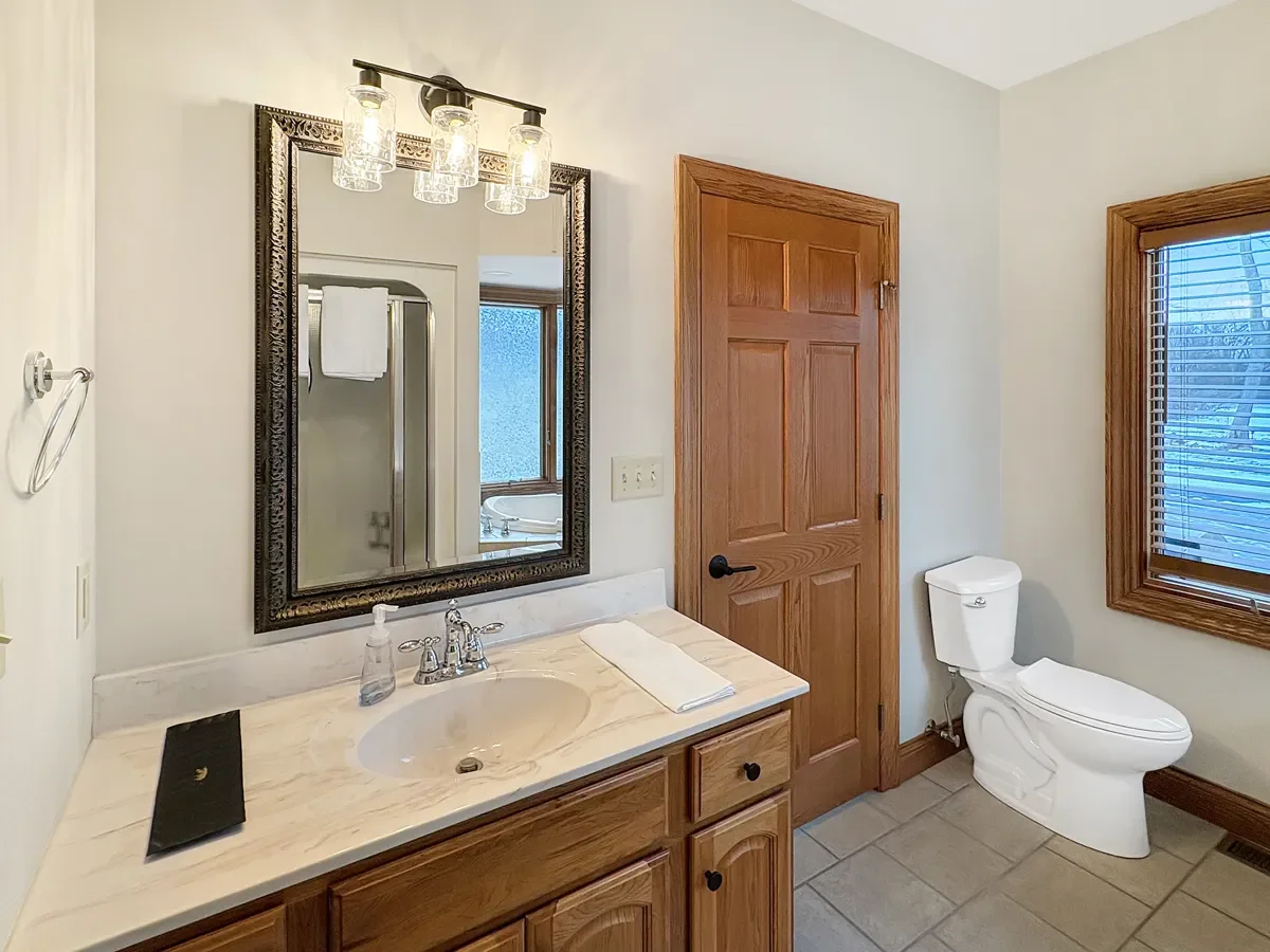 Bathroom with wooden door, window with blinds, white toilet, granite countertop with sink, mirror, wall-mounted light fixture, and towel.