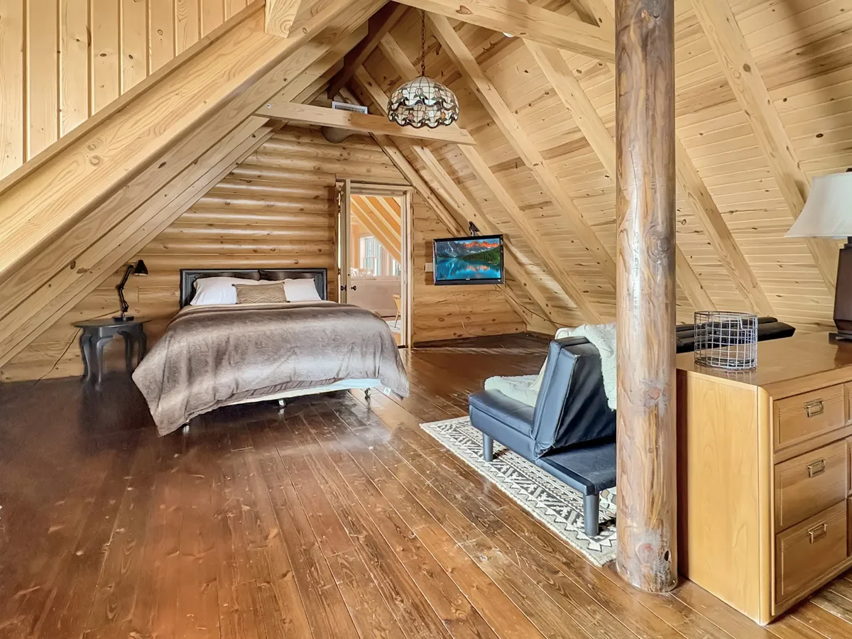 A cozy attic bedroom with wooden walls, a bed with beige and white linens, a bedside table with a black lamp, a small sofa, a wall-mounted TV, and a dresser with a lamp on it.