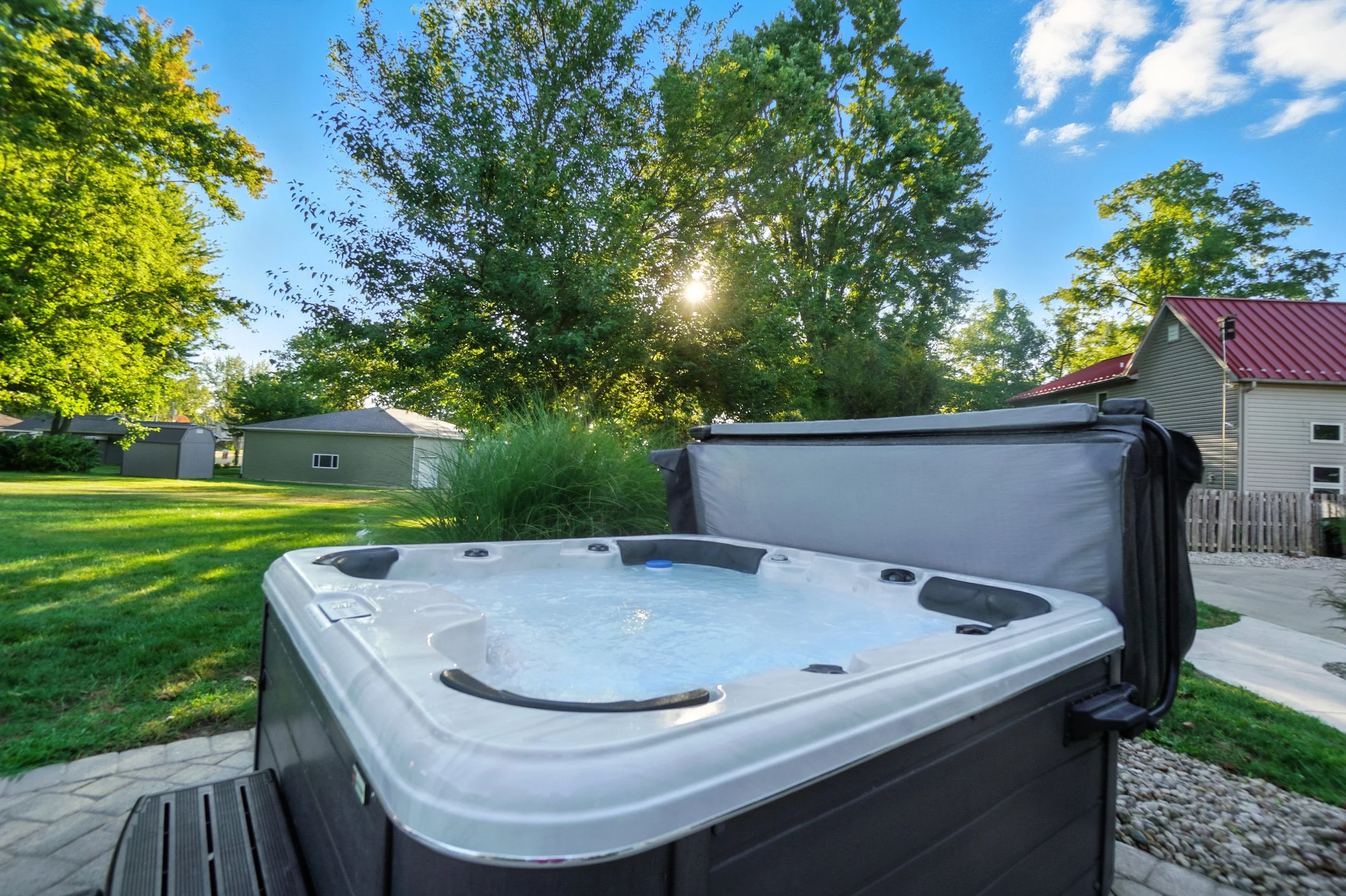A hot tub outdoors in a backyard with green grass, trees, and a partly cloudy blue sky.