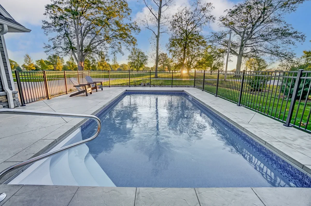 Empty outdoor swimming pool with a safety fence, two wooden lounge chairs, and a scenic view of trees and sunset.