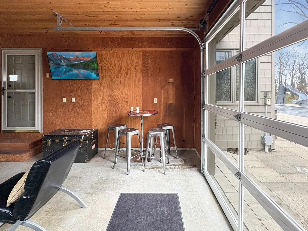 A garage converted into a game or lounge area with a mounted TV, a small round table with three metal stools, and a vintage trunk. The open garage door reveals an outdoor patio and yard.