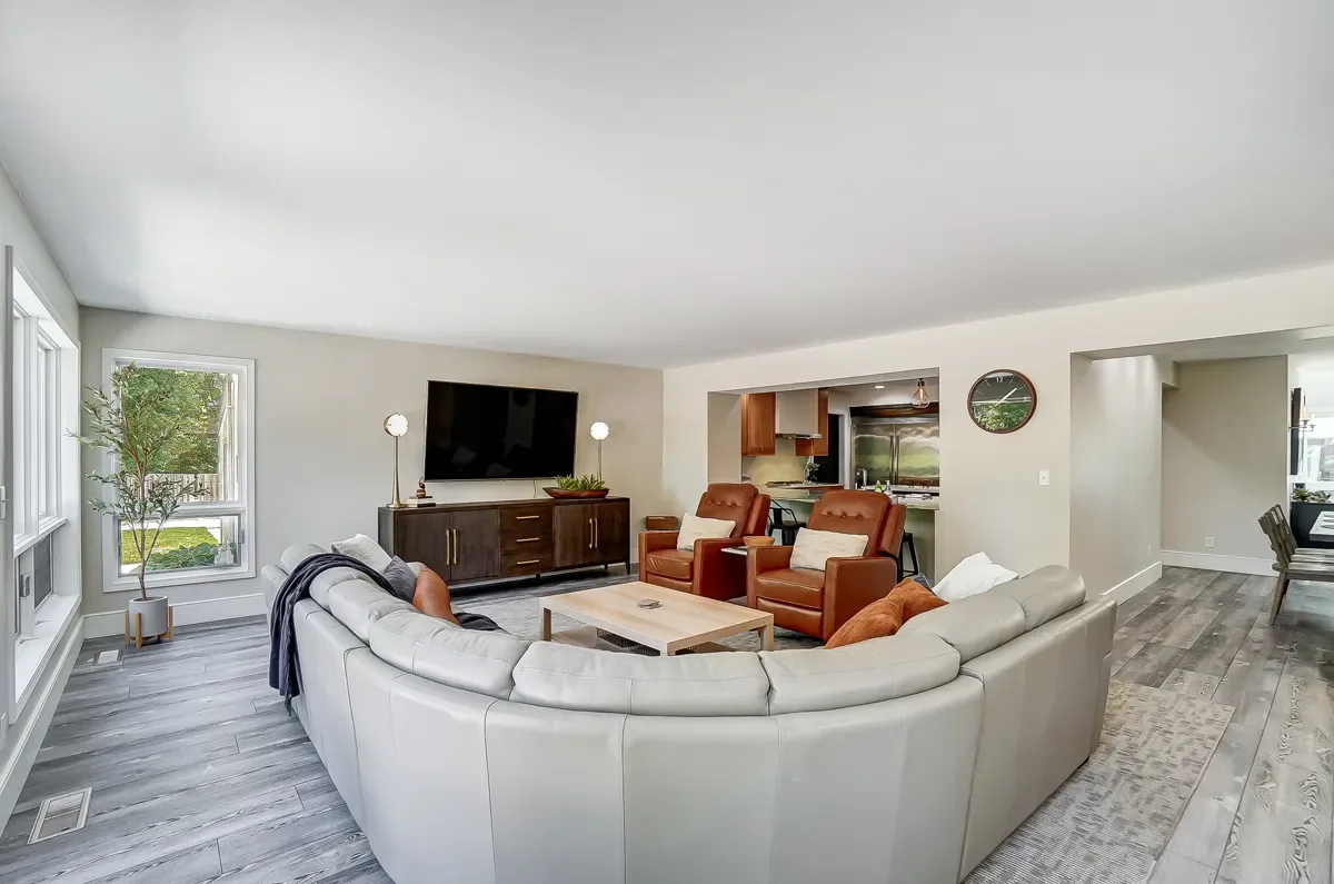 Living room with curved white sofa, two brown armchairs, a wooden coffee table, and a large TV on a dark wood stand, with a kitchen visible in the background.