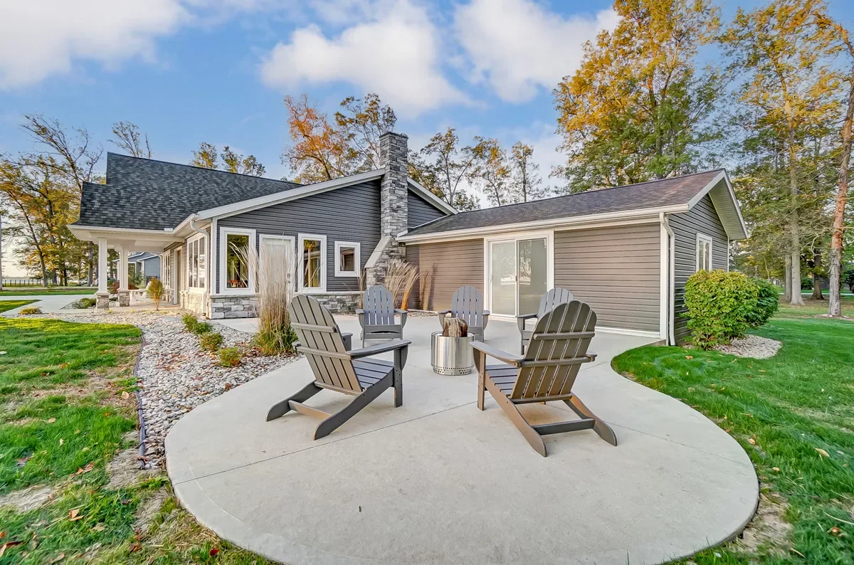 Backyard with concrete patio and four Adirondack chairs around a small fire pit, house with dark gray and beige siding, large trees, and a partly cloudy sky.