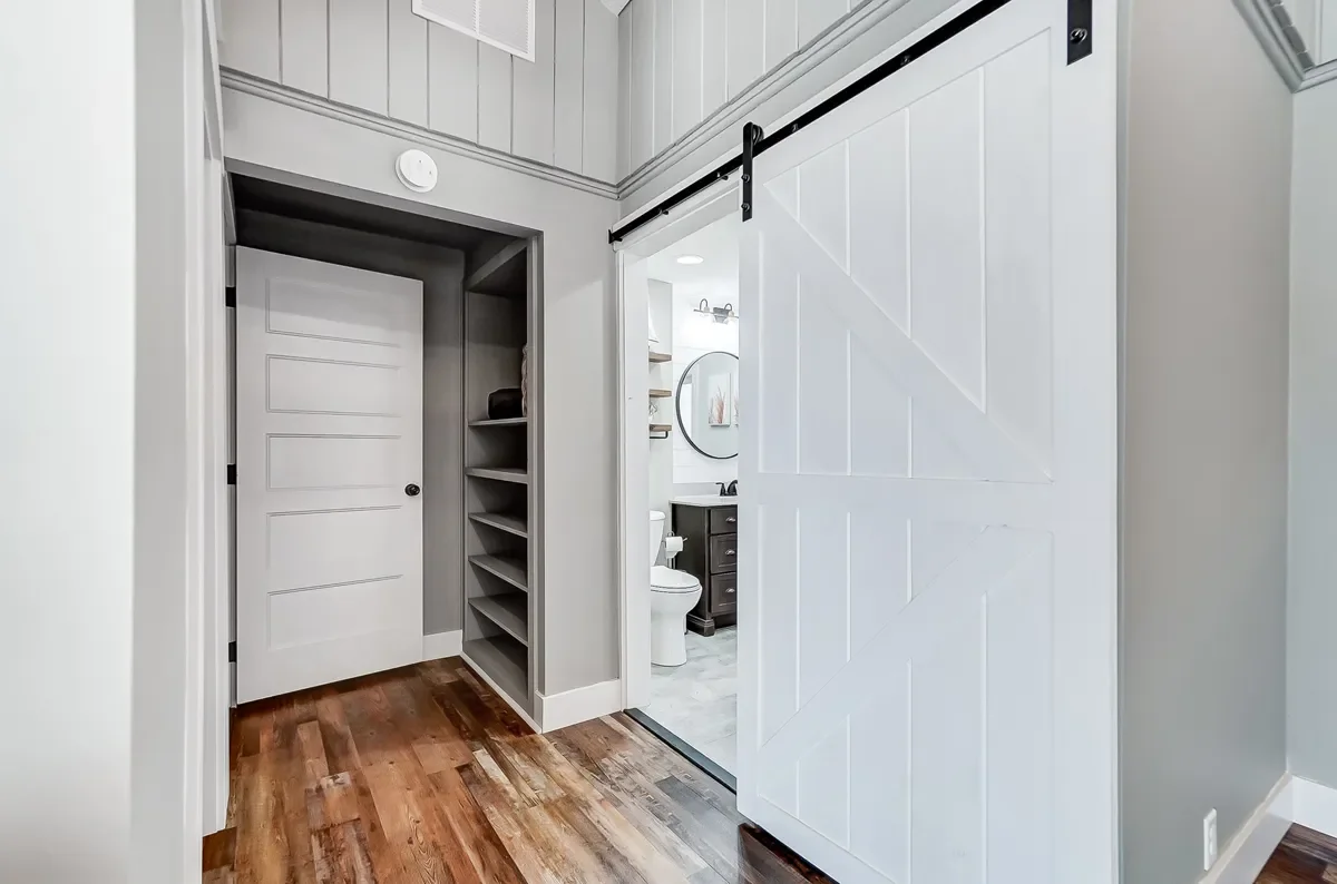 Interior view showing a sliding barn door leading to a bathroom with a toilet, sink, and mirror, and a small room with shelves and a closed door, hardwood floors, and light gray walls.