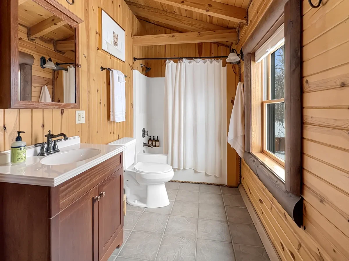 Bathroom with log cabin style wood paneling, a vanity with a mirror, a white toilet, a bathtub with a shower curtain, a window, and a framed picture of a cat.