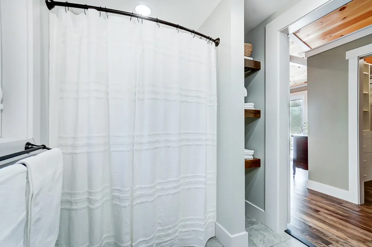 Bathroom with a white shower curtain, towels on a towel rack, open shelves with folded towels and a wicker basket, and a view of a room with wooden ceiling and floors.