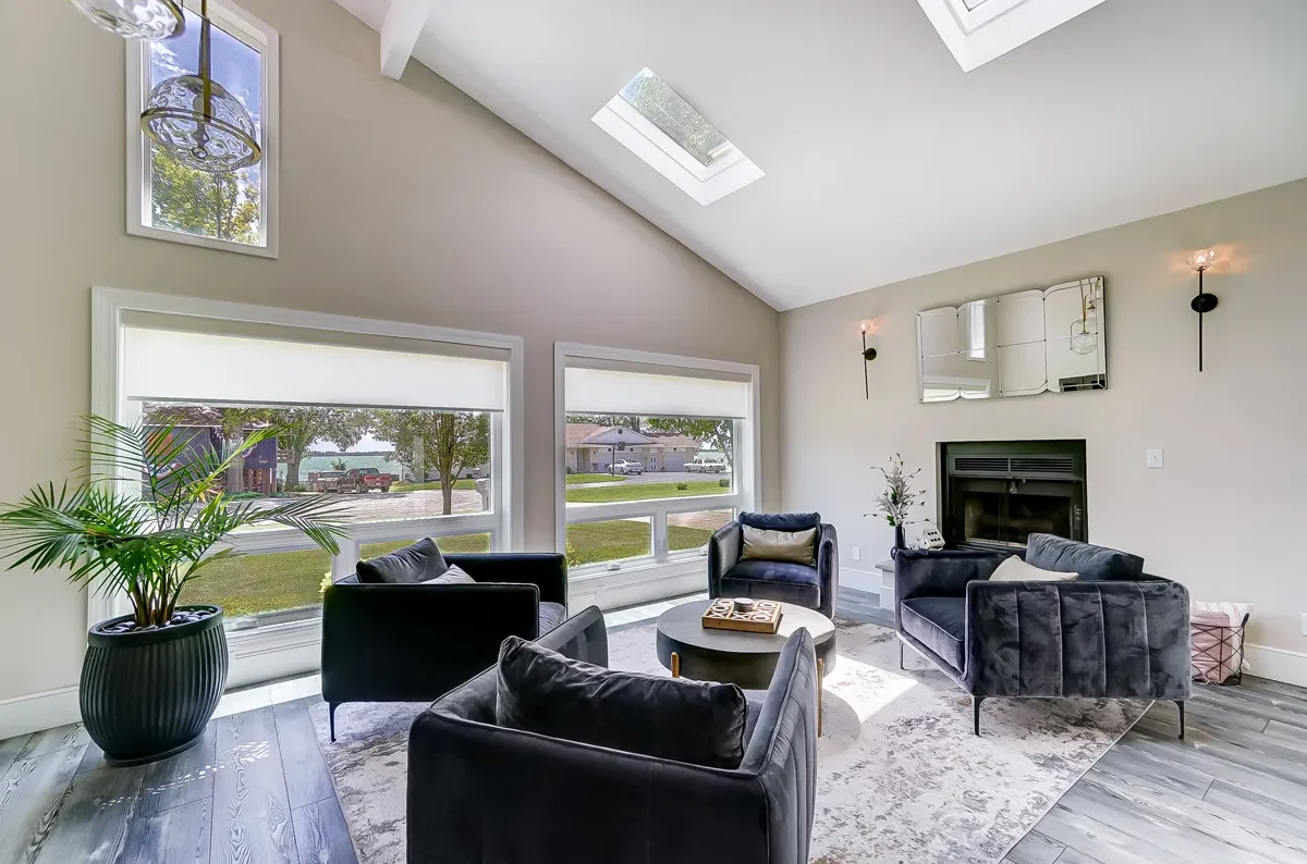 Bright living room with large windows, skylights, modern black armchairs, a white round coffee table, a fireplace, a mirror above the fireplace, wall sconces, and a potted plant on the left.