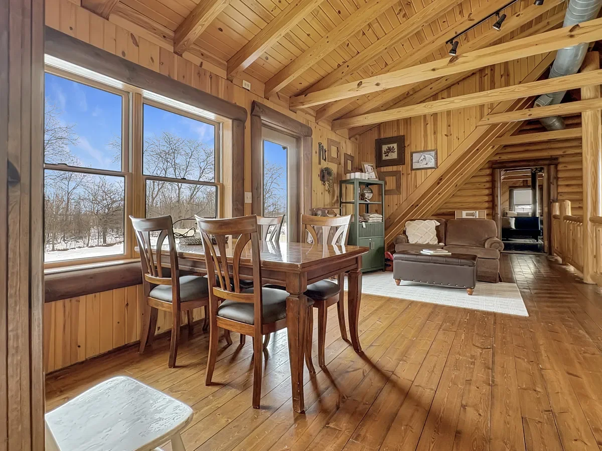 Interior of a cozy cabin with wooden walls and ceiling, featuring a dining table with chairs, a large window showing a snowy landscape outside, and a small living area with a gray couch, a white rug, and wall art.