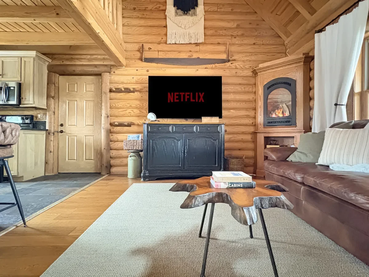 Living room in a log cabin-style interior with a dark TV displaying the Netflix logo on a black cabinet, a leather sofa with pillows, a rustic coffee table with books, a fireplace inset in the wall, and kitchen area visible to the left.