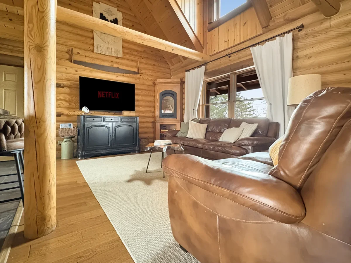 Cozy cabin living room with wooden walls, leather sofas, a flat-screen TV displaying Netflix, and large windows with white curtains.