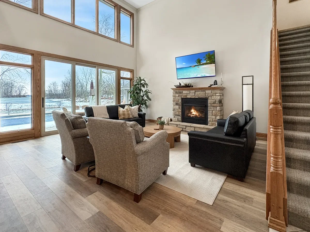 Living room with large windows showing snowy outdoors, a fireplace with a flat-screen TV above it, and three sofas arranged around a wooden coffee table.
