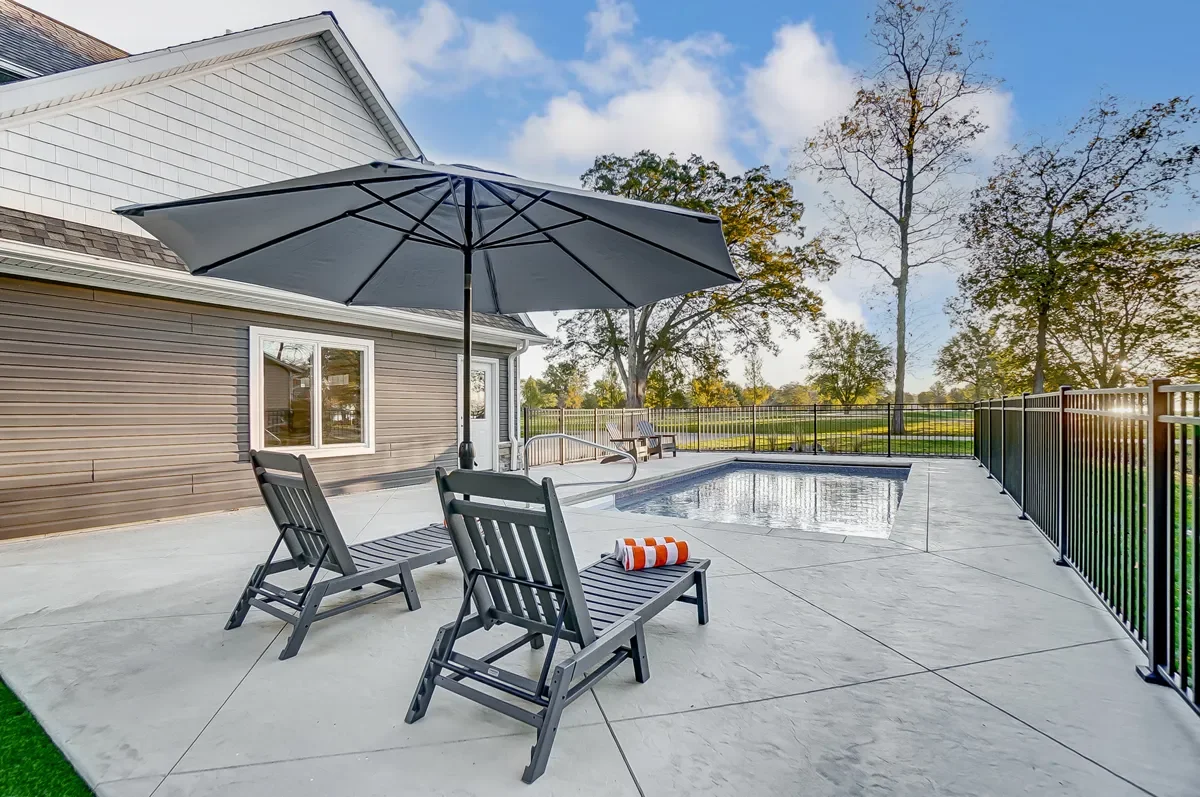 Residential backyard with a swimming pool, patio chairs, a large umbrella, and a view of trees and a park in the background.