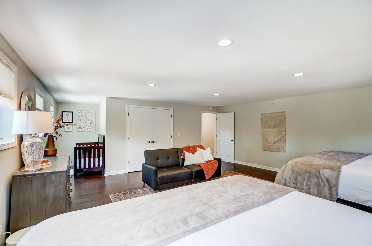 A spacious bedroom with two beds, a black loveseat with white and orange pillows, a dresser with a lamp and decorative items, and a small area with a crib near the window. The walls are light-colored and there are framed pictures and artwork hanging.