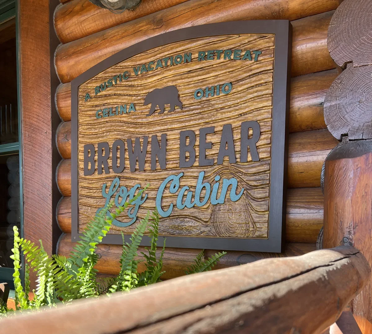Wooden sign reading "A rustic vacation retreat Ohio Celina Brown Bear Log Cabin" with a bear silhouette, mounted on a log cabin wall, with green ferns in front.