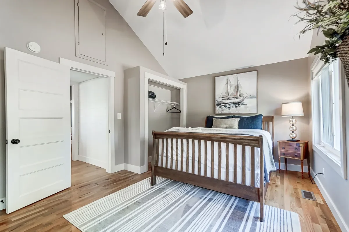 A bedroom with a wooden bed frame, white linens, and pillows. There's a bedside table with a lamp, a painting of a sailboat on the wall, and a window with blinds. The floor is hardwood with a striped rug, and there's an open closet with a hanger. The