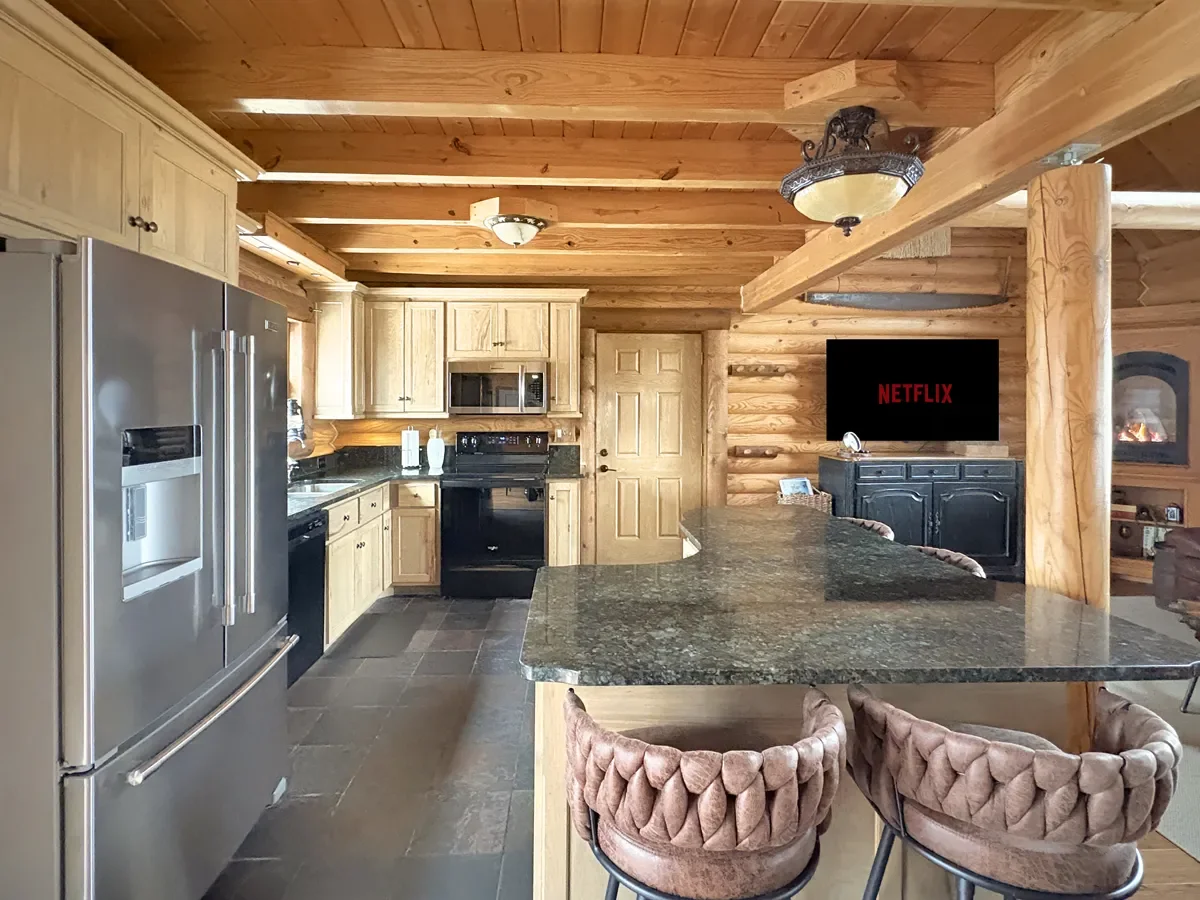 A rustic kitchen with wooden walls, ceiling, and cabinetry, featuring stainless steel appliances, a black stove, and dark granite countertops. There’s a large kitchen island with barstools, and a door in the background. To the right, a small living a