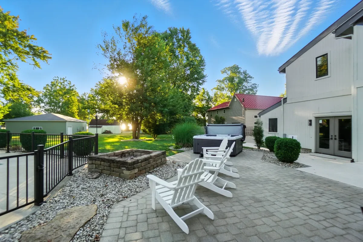 Backyard patio with Adirondack chairs and a hot tub, enclosed by a black metal fence, with trees, shrubs, and neighboring houses in the background.