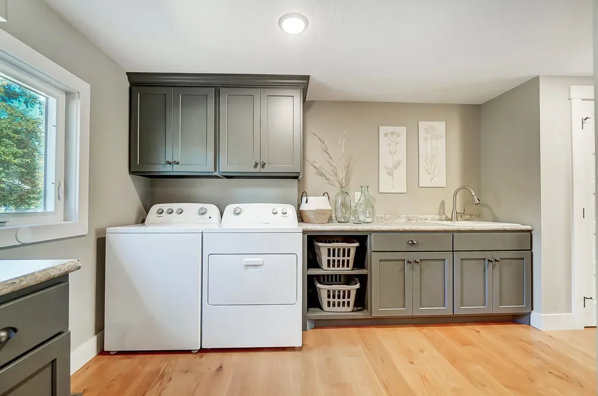 Laundry room with a washing machine, a dryer, gray cabinets, a window, decorative vases, and framed artwork on the wall.
