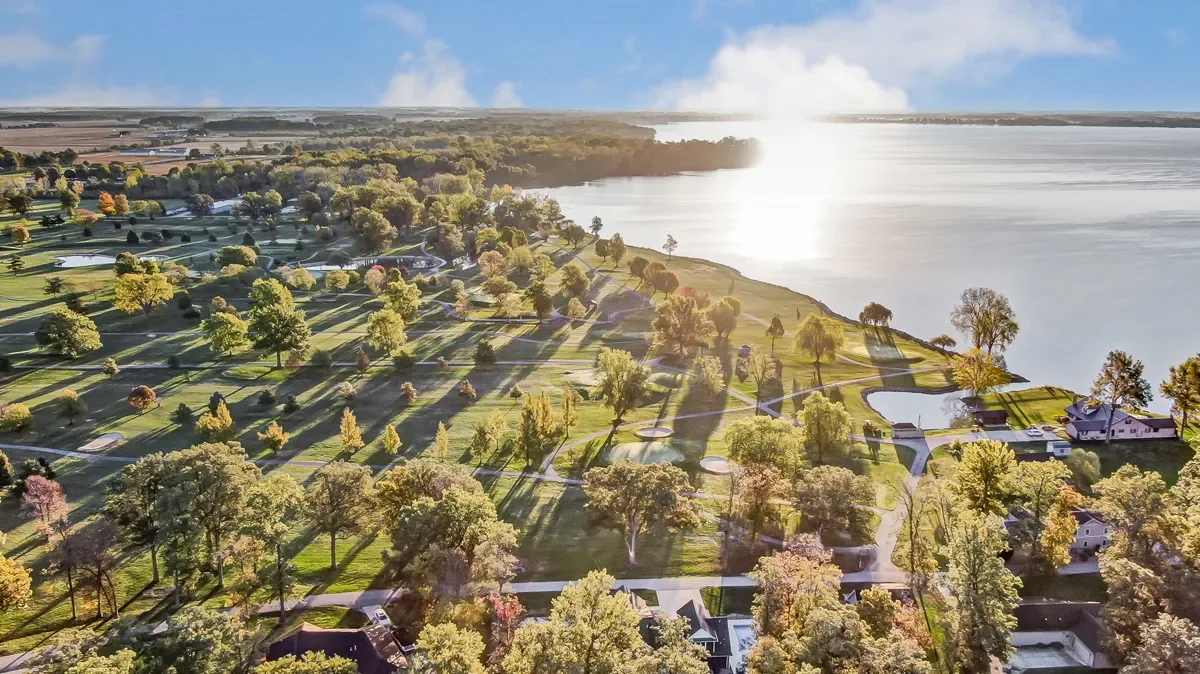 Aerial view of a golf course near a large body of water, with trees, houses, and pathways.