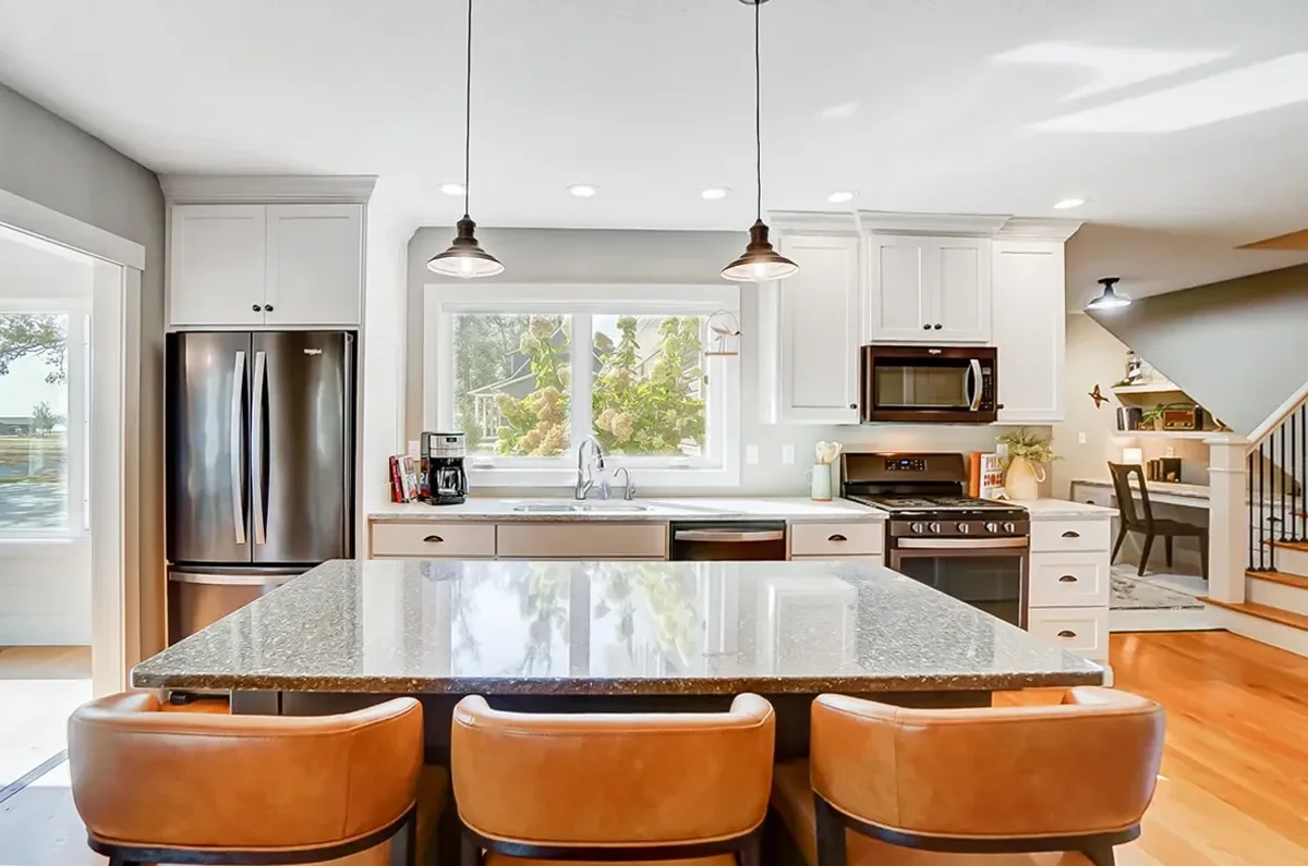 Modern kitchen with stainless steel refrigerator, white cabinets, granite island, and three pendant lights, with a window above the sink showing trees outside.