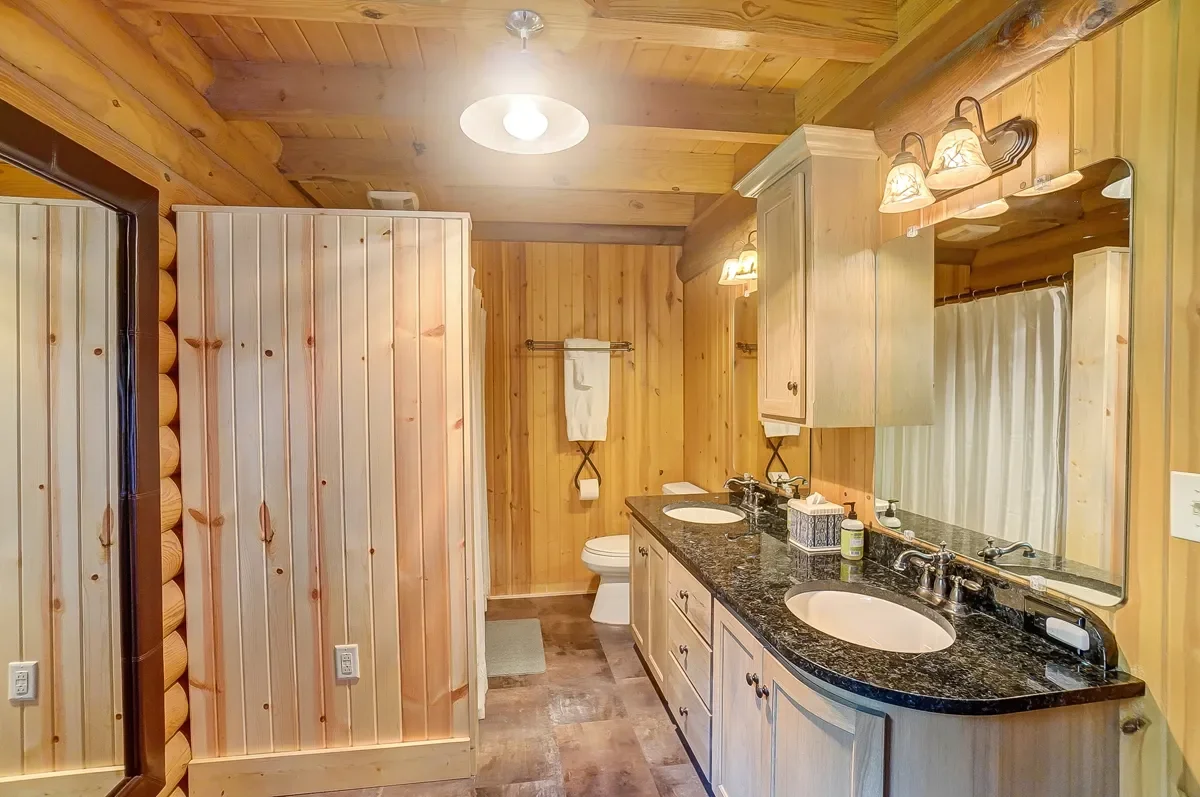 Wood-paneled bathroom with dual sinks, granite countertop, mirror, toilet, towel rack, and warm lighting.