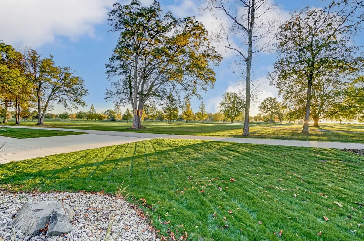 A park with green grass, trees, and a paved pathway, with the sun setting in the background.