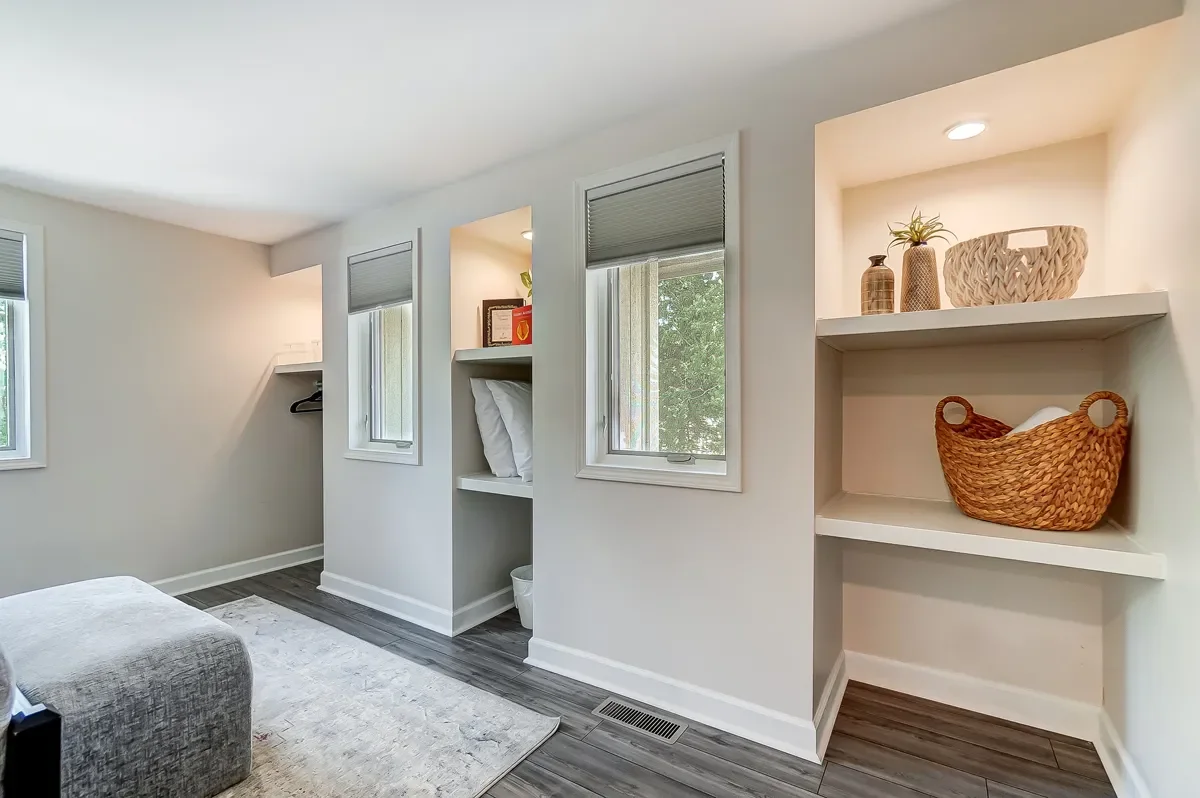 A corner of a bedroom with a bed, a gray ottoman, and built-in wall shelves with decor and windows with blinds.