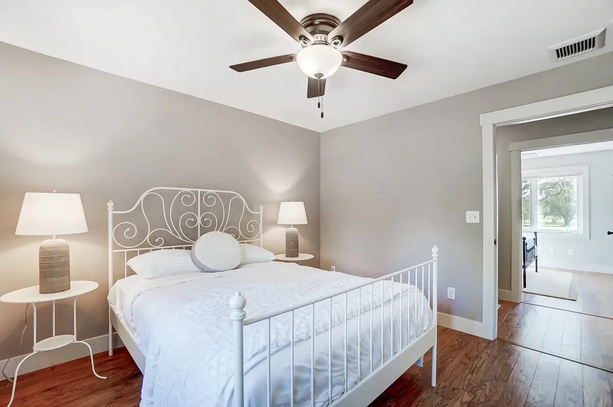 Bedroom with white metal bed, two matching nightstands with lamps, gray walls, hardwood floors, ceiling fan, doorway to another room with window and dark bed frame.