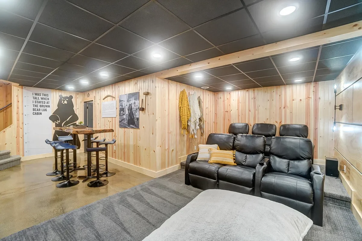 A cozy basement room with wood-paneled walls and a dark ceiling. It features a black leather recliner sofa with throw pillows, a small gray rug, and a beige ottoman. There is a high-top table with four barstools near a wall decorated with a bear mura