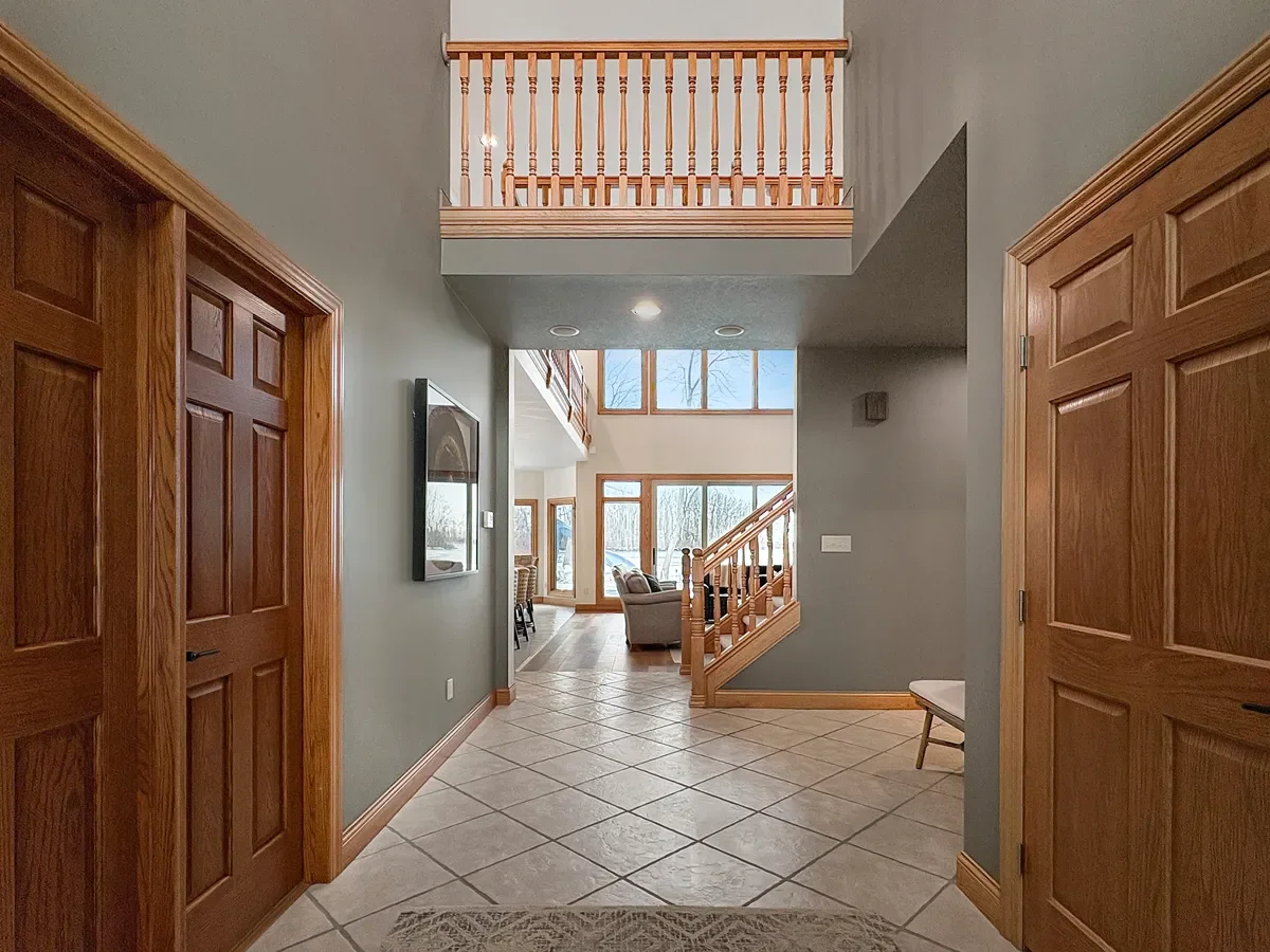 Interior view of a house foyer with tiled floor, wooden doors, and a staircase leading to a second level with a wooden railing, large windows allowing natural light, and a view of the living area with couches.