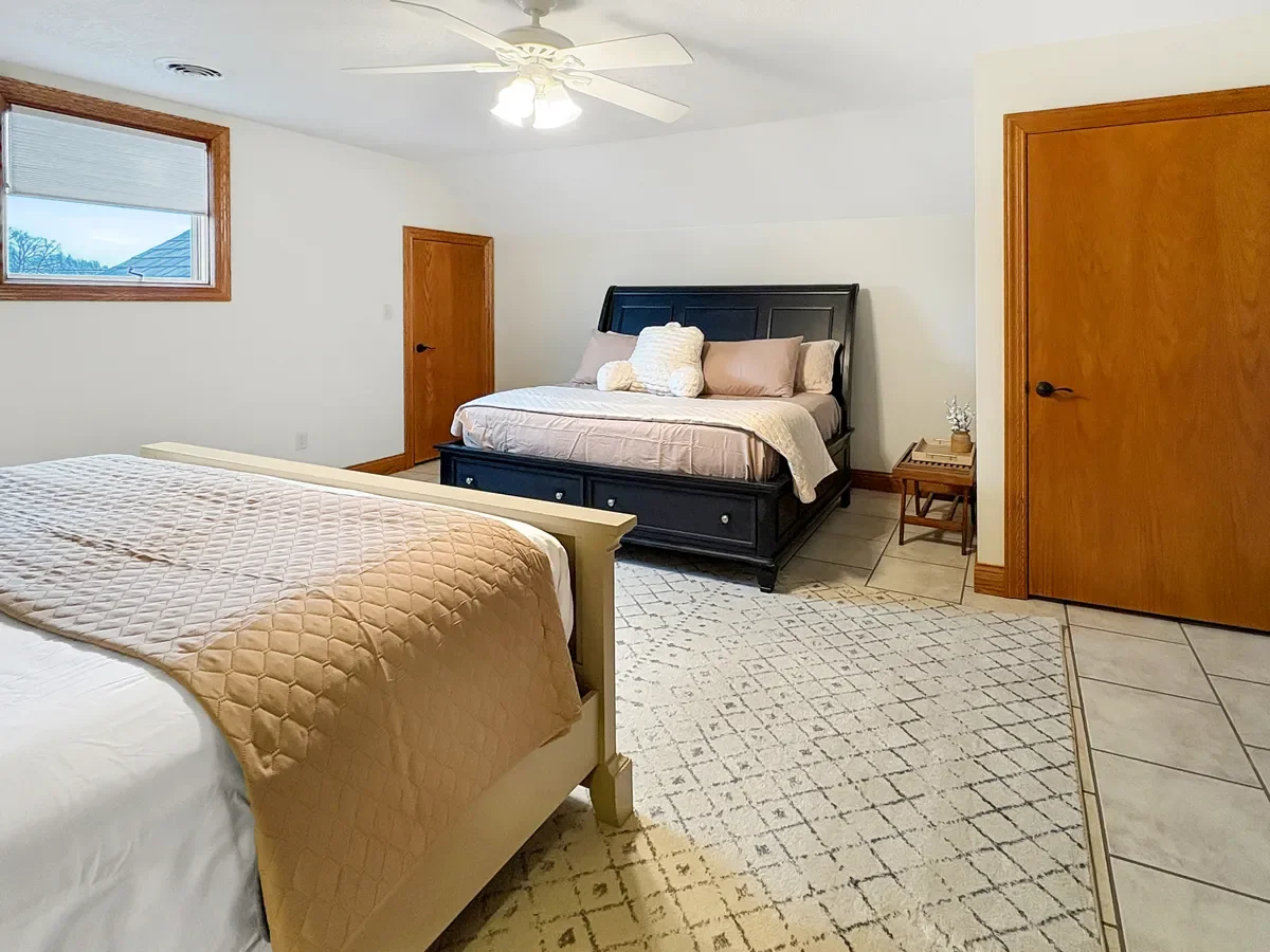 Bedroom with two beds, one with a beige quilt and the other with pillows and a stuffed animal, a ceiling fan, window with a blind, wooden doors, and a small side table with a decorative object.